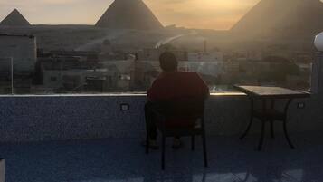Una persona sentada en una silla observando una vista urbana con pirámides al fondo durante el atardecer.