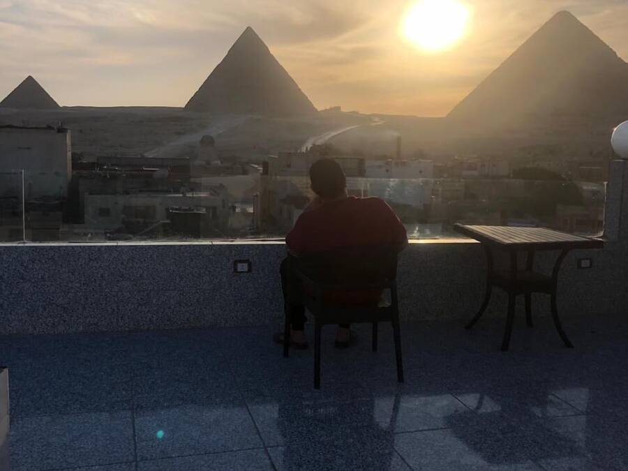 Una persona sentada en una silla observando una vista urbana con pirámides al fondo durante el atardecer.