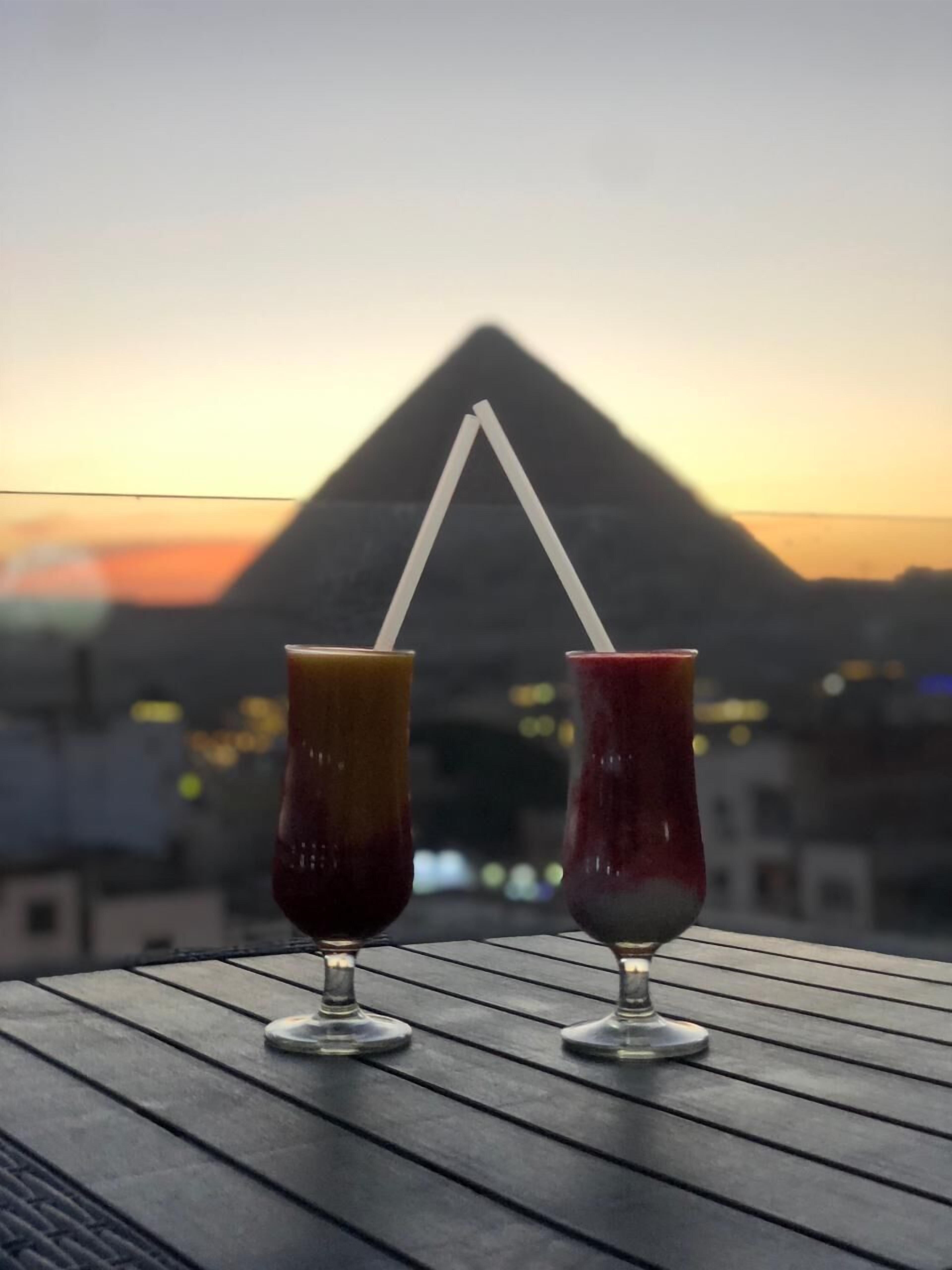 Duas bebidas com canudos sobre uma mesa, com vista para uma pirâmide ao fundo.