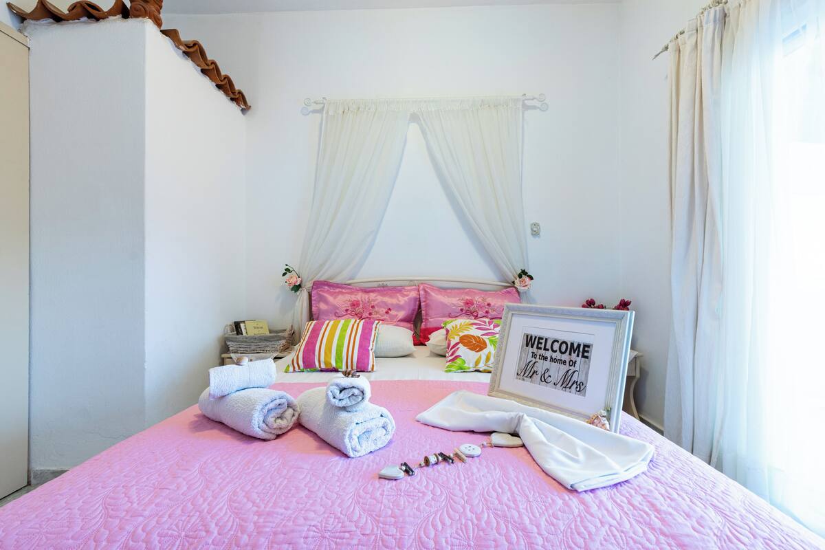 A bedroom with a pink quilted bedspread, neatly folded towels, a welcome sign, and a decorative pillow arrangement.