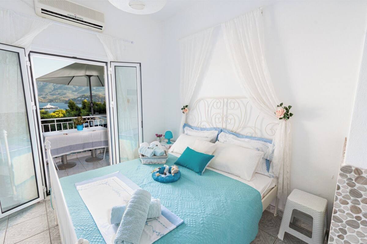 A bedroom with a bed, a bedside table, a small stool, a window with a view of the sea, and a balcony with a table and chairs.