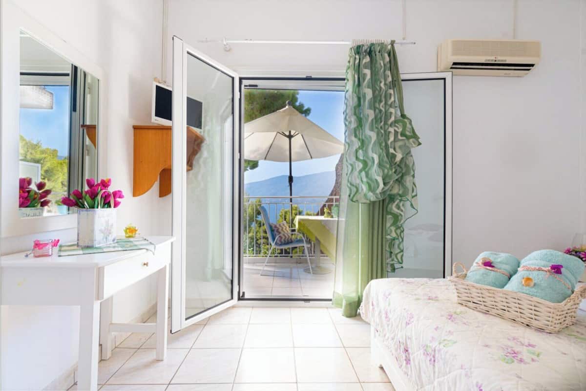 A bedroom with a balcony, a white desk, a bed with a floral bedspread, and a wicker basket with towels.