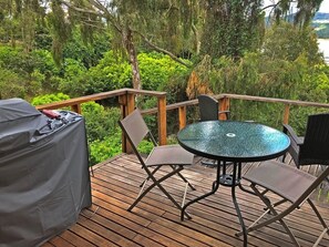 Una terraza de madera con una mesa y sillas de vidrio, rodeada de abundante vegetación.