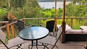 Une terrasse en bois avec une table et des chaises en verre, donnant sur un lac où l’on aperçoit des bateaux et, au loin, des collines.