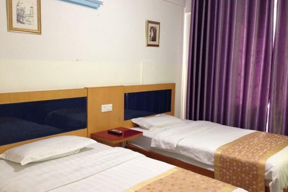 A hotel room with two single beds, a wooden headboard, a small bedside table with a book, and a window with purple curtains.