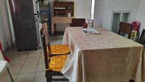 A dining area with a table covered by a beige tablecloth, two chairs with yellow cushions, a refrigerator, a shelf with dishes, and a window with natural light.