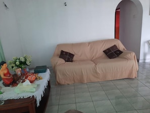 A living room with a beige sofa, a small table with religious statues and offerings, and a tiled floor.