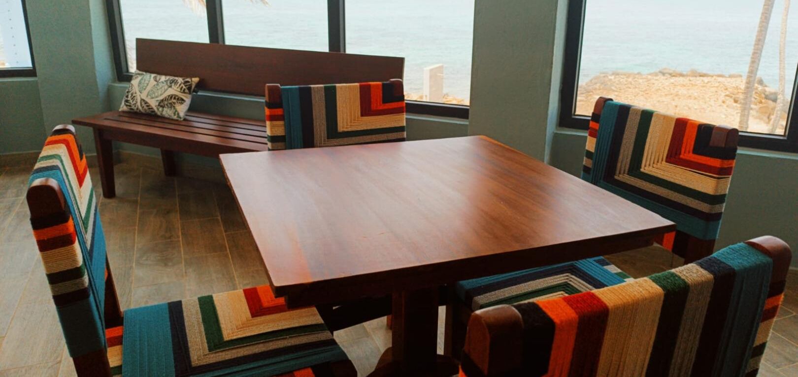 A room with a wooden table and chairs, a bench, and large windows with a view of the beach.
