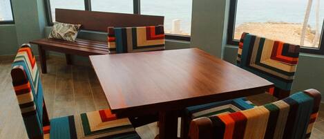 A room with a wooden table and chairs, a bench, and large windows with a view of the beach.