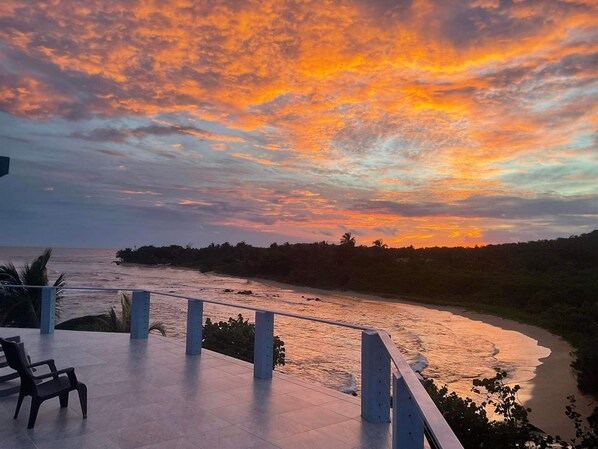 A sunset view from a terrace with a railing overlooking a river and a distant shoreline. - Mama Lola Bed and Breakfast (Corn Islands)
