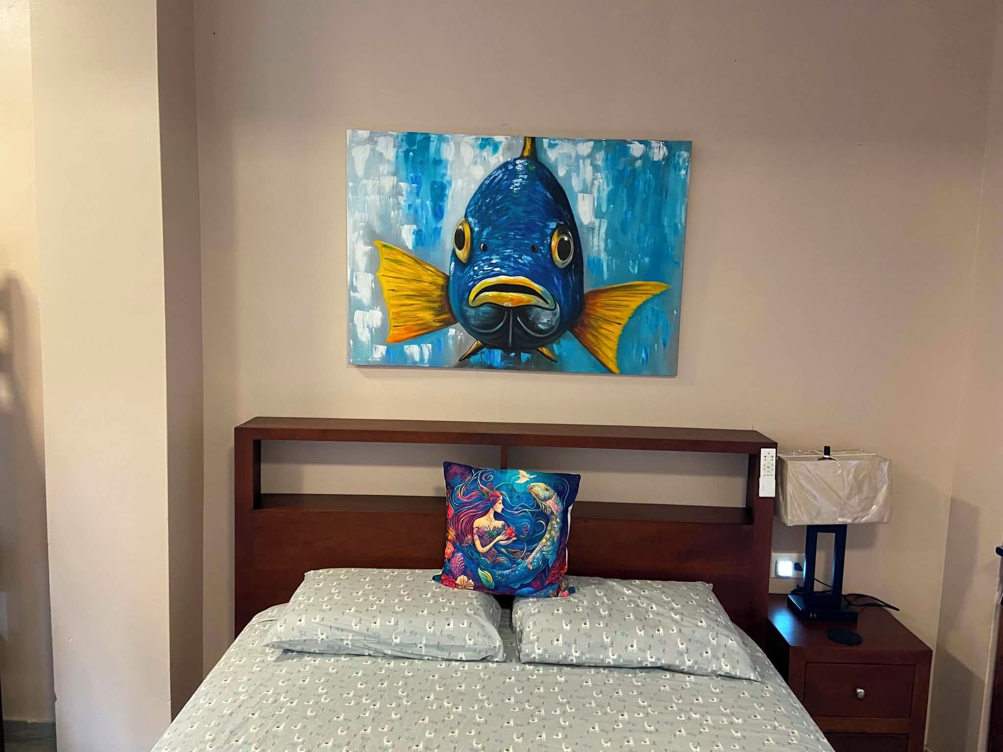 Un dormitorio con cama, almohada, mesita de noche, lámpara y un cuadro de un pez en la pared.