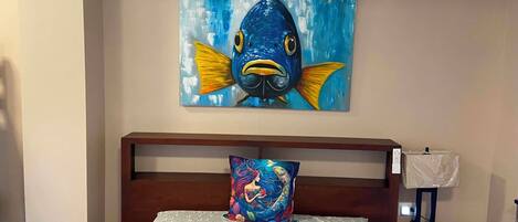 Un dormitorio con cama, almohada, mesita de noche, lámpara y un cuadro de un pez en la pared.