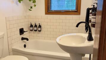 Une salle de bain avec une baignoire blanche, un lavabo blanc et des toilettes. Il y a une fenêtre avec des stores et une plante suspendue.