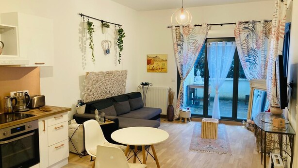 Et moderne køkken med hvide skabe og en indbygget ovn, en grå sofa med dekorativt sengegavl, et lille rundt bord med stole, samt et stort vindue med gardiner.