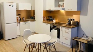 Et lille køkken med hvide skabe, indbygget ovn, vask og spiseplads med to stole.