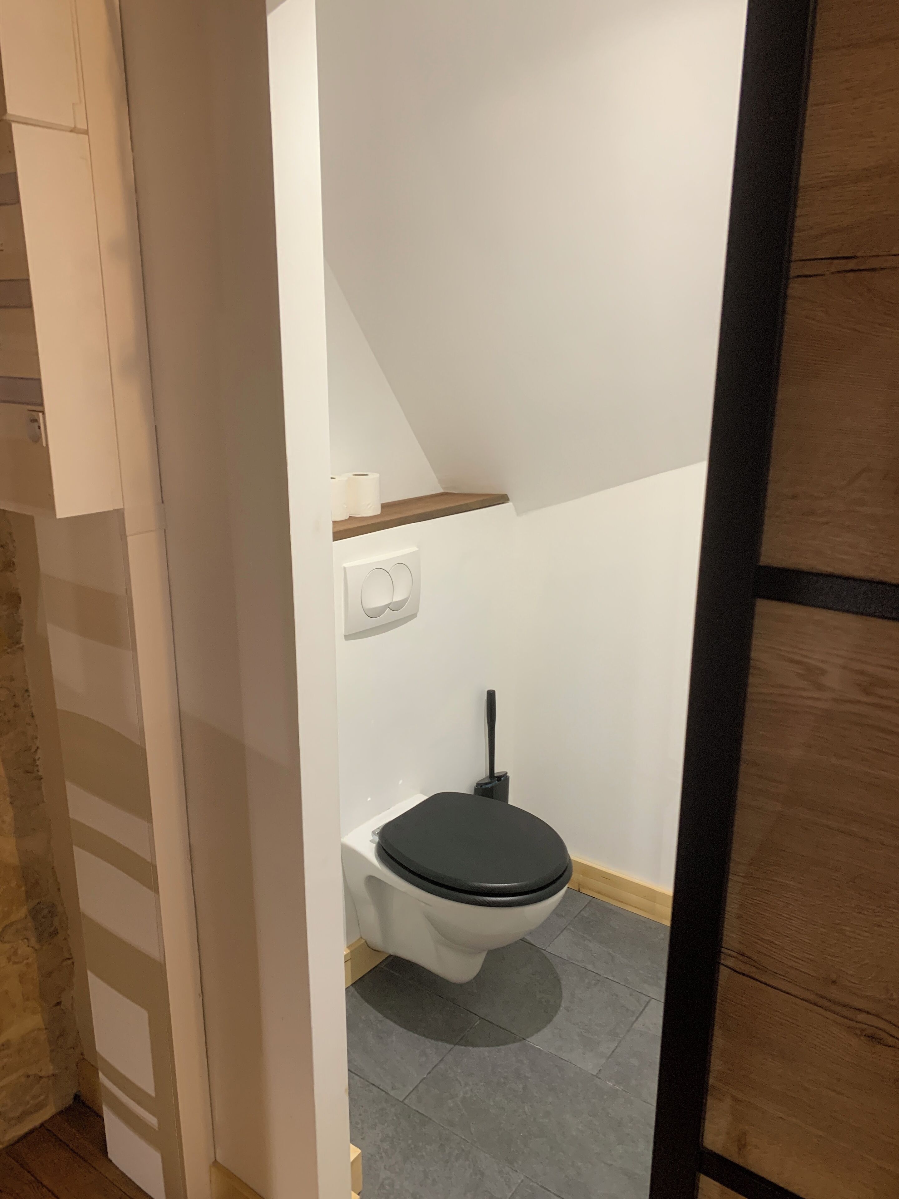 A small, modern bathroom with a white toilet, a wooden shelf, and a tiled floor.