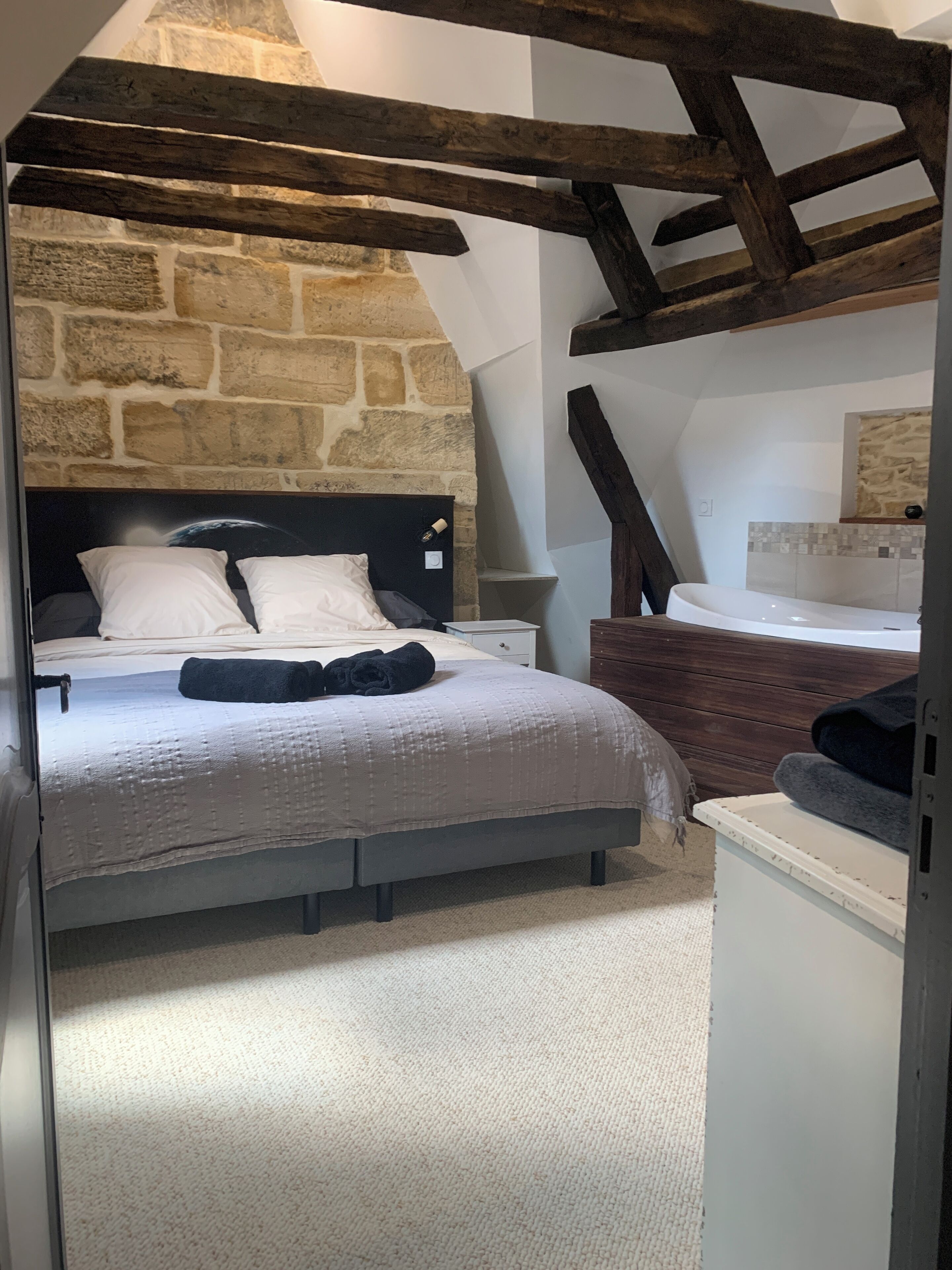 Une chambre avec un mur en pierre, un lit avec une tête de lit, un banc en bois et une table de chevet blanche.