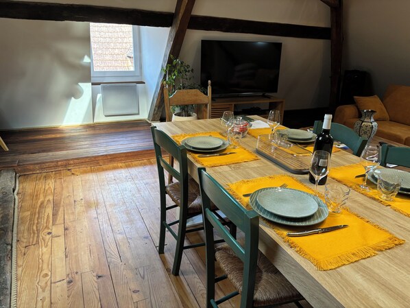 A dining area with a wooden table set for four, a television in the background, and a window with a view of the outside.