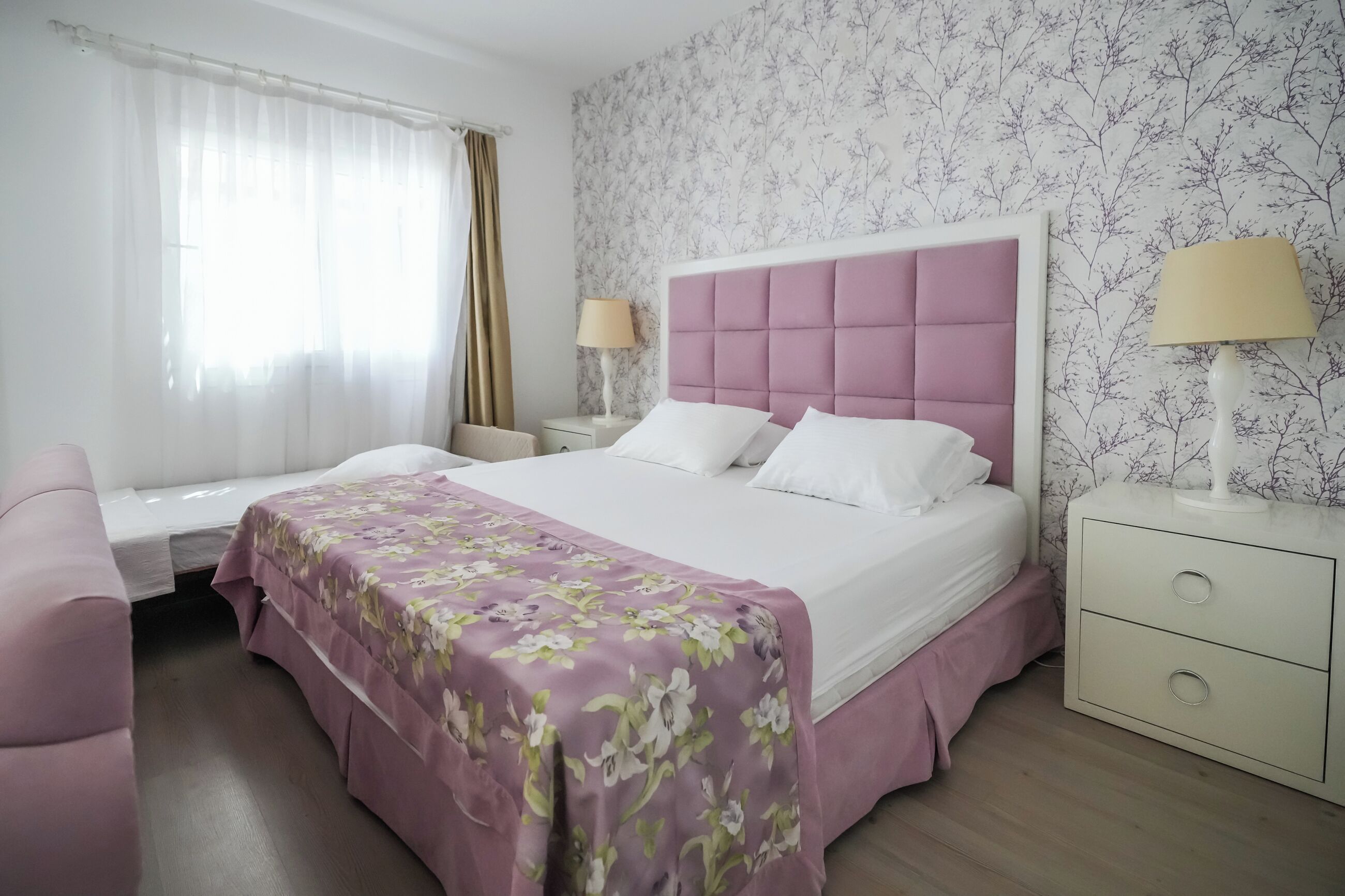 Una camera d'albergo con un letto, un comodino, una lampada, una sedia e una finestra con tende.