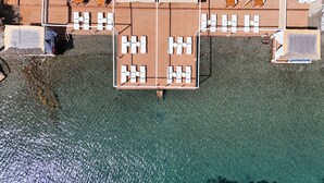 Eine Vogelperspektive zeigt eine Strandterrasse mit mehreren Liegestühlen und einem kleinen Tisch, umgeben von klarem, türkisblauem Wasser.