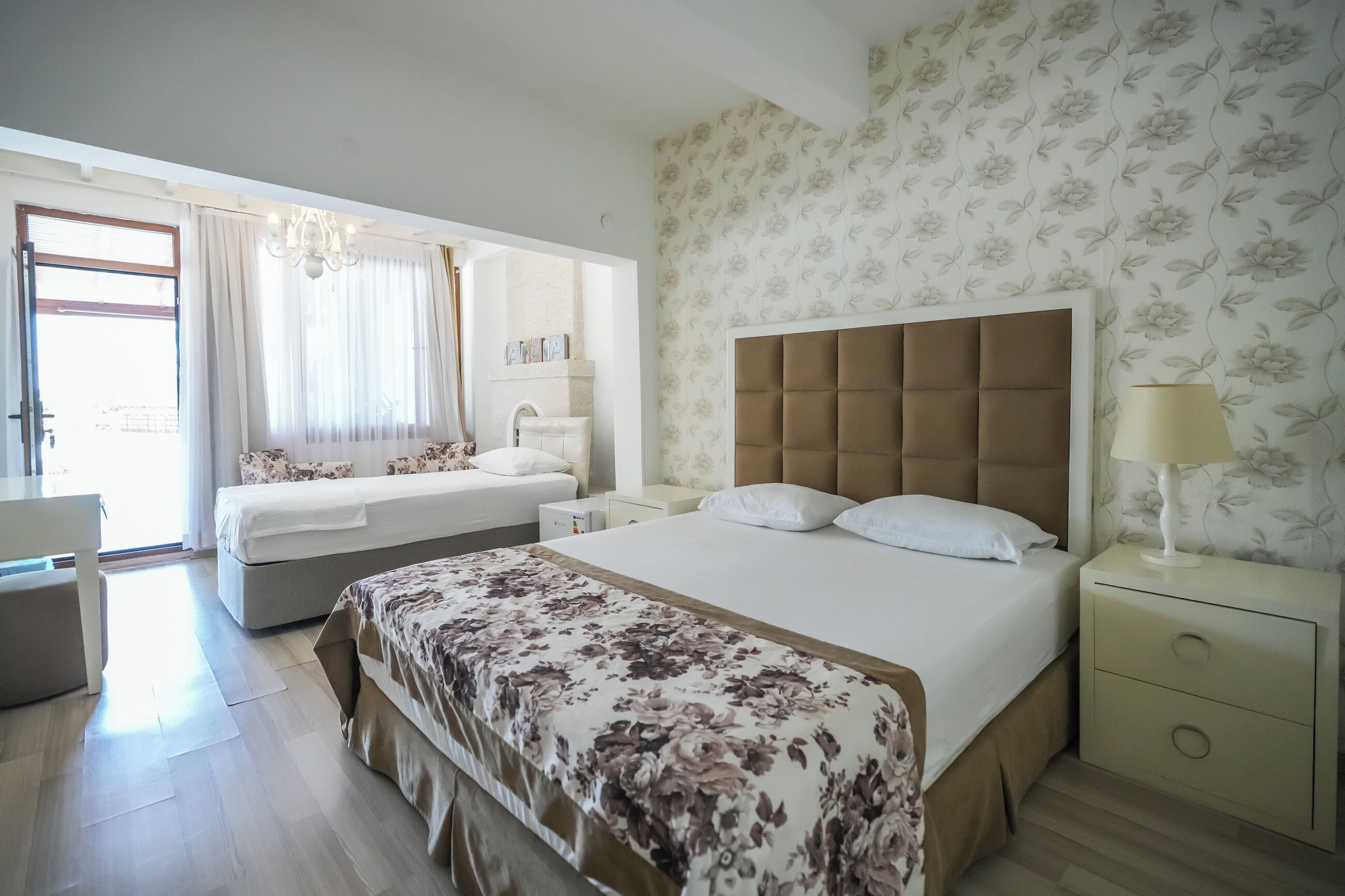 A hotel room with two beds, a large window with curtains, a chandelier, and a floral wallpaper.