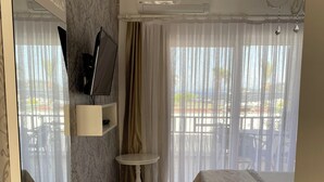 Ein Hotelzimmer mit Bett, einem kleinen runden Tisch, einem an der Wand montierten Fernseher und Blick ins Freie durch transparente Vorhänge.