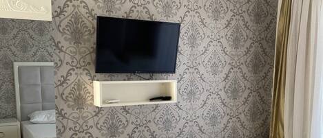 Un dormitorio con un televisor montado en la pared, una mesita de noche, una cama con colcha estampada y una mesita con lámpara.