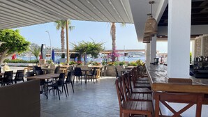 Ein Außenbereich mit Tischen und Stühlen, Blick auf das Meer und ein überdachter Bereich mit einer Bar.