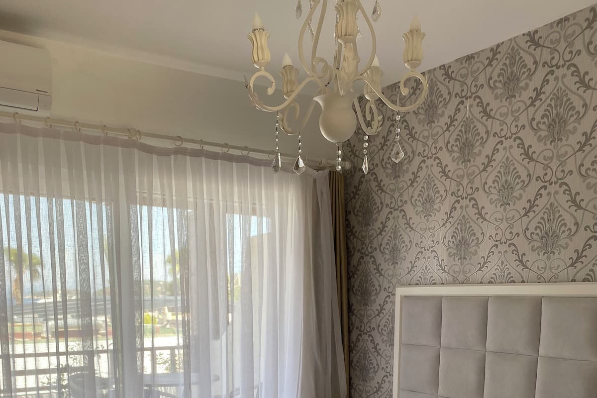 A spacious bedroom with a large bed, a chandelier, and a view of the outdoors through sheer curtains.