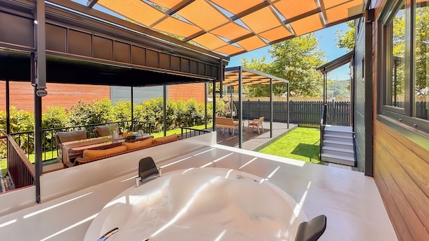 A modern outdoor hot tub area with a pergola overhead, a wooden deck, and a view of a backyard with a lawn and a wooden fence. - Baronet Tiny House Otel (Yalova)