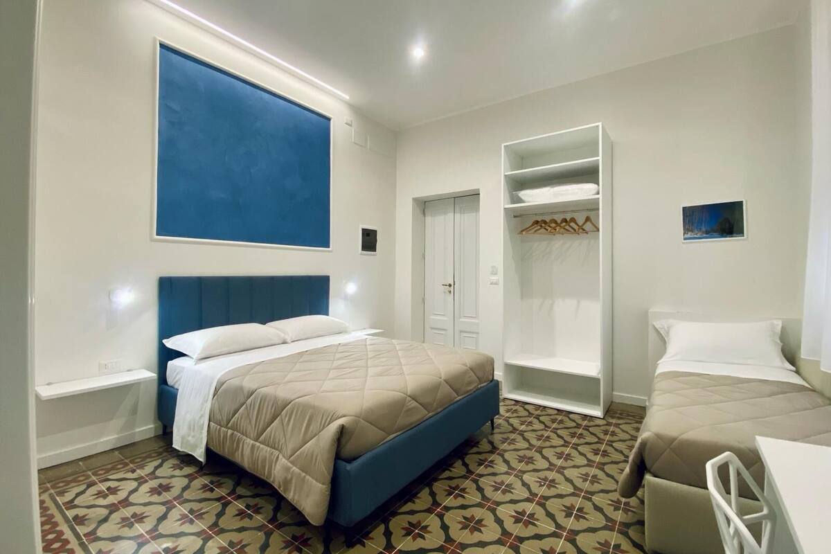 A hotel room with two beds, a wardrobe, a large blue artwork on the wall, and patterned floor tiles.