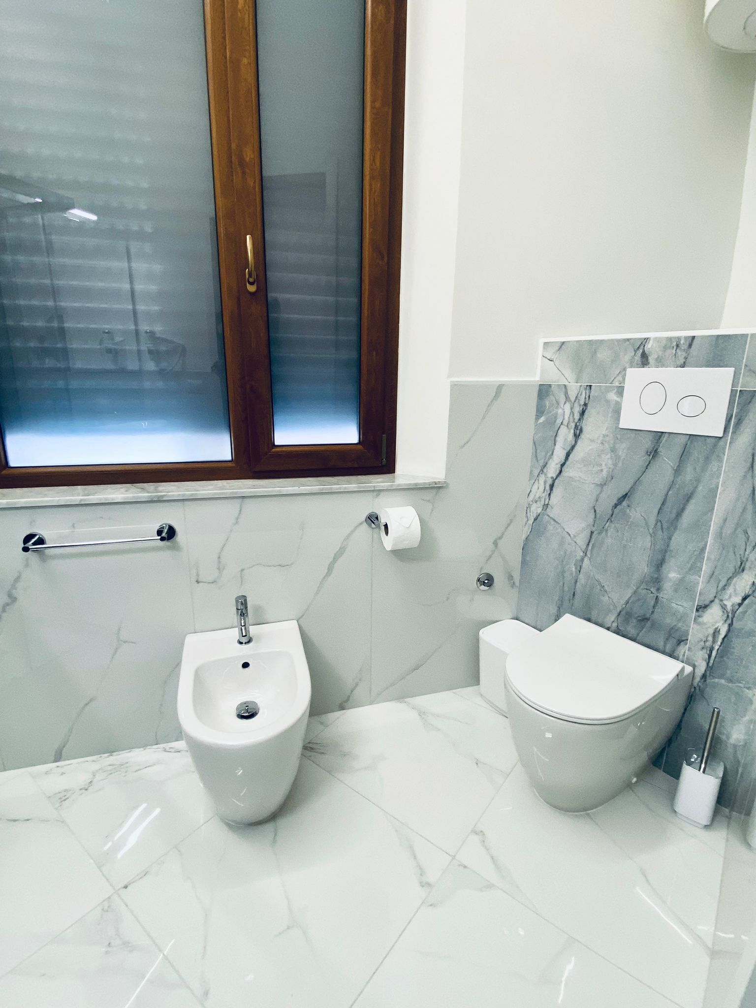 A bathroom with a bidet, toilet, marble tiles, a window with frosted glass, and a towel rack.