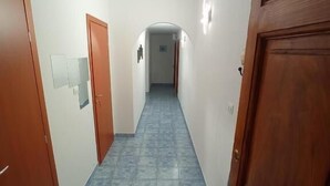 A hallway with blue-tiled flooring, wooden doors on both sides, and a white wall with a light switch and a small picture. - Aria di Sale Hostel (Trieste)