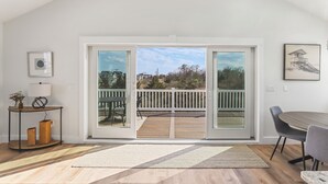 Ein Zimmer mit einer großen Schiebeglastür, die auf eine Terrasse mit Holzgeländer führt, einem Esstisch mit Stühlen und einem kleinen Konsolentisch mit Lampe und Dekoration.