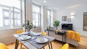 A modern dining area with a table set for two, a sofa, a TV, and a window with a city view.