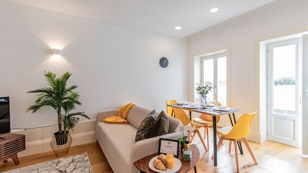 A modern living room with a sofa, dining table, and chairs. There is a plant, a clock, and a framed welcome sign. - Rodrigues de Freitas 391 2J by LovelyStay (Porto)
