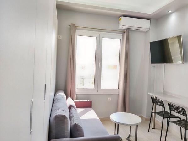 A modern living room with a sofa, a small round table, two chairs, a TV mounted on the wall, and a window with curtains.