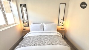 Un lit bien fait, avec une literie blanche et un couvre-lit à motifs, flanqué de deux tables de chevet avec des lampes, dans une pièce dotée d’une fenêtre et d’un panneau décoratif fixé au mur.
