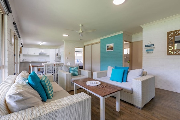 A modern, well-lit living area with wicker furniture, a wooden coffee table, and a kitchen in the background. - Bella Rose - 3 BR w/ Boat Parking - Walk to Beach (Agnes Water)