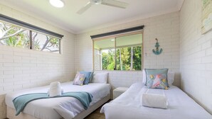 A room with two beds, white brick walls, a ceiling fan, and large windows with a view of greenery.