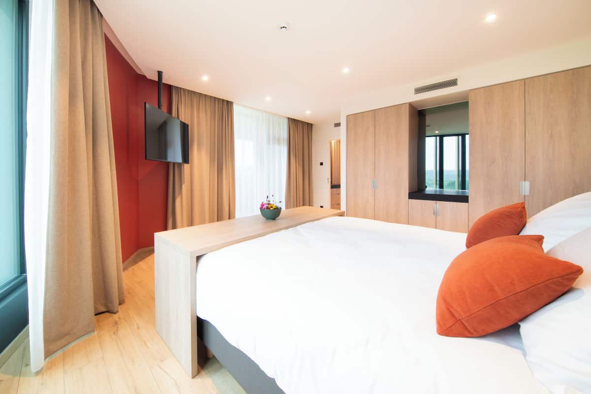 A modern bedroom with a large bed, wooden headboard, a flat-screen TV mounted on the wall, and a bedside table with a bowl of flowers.