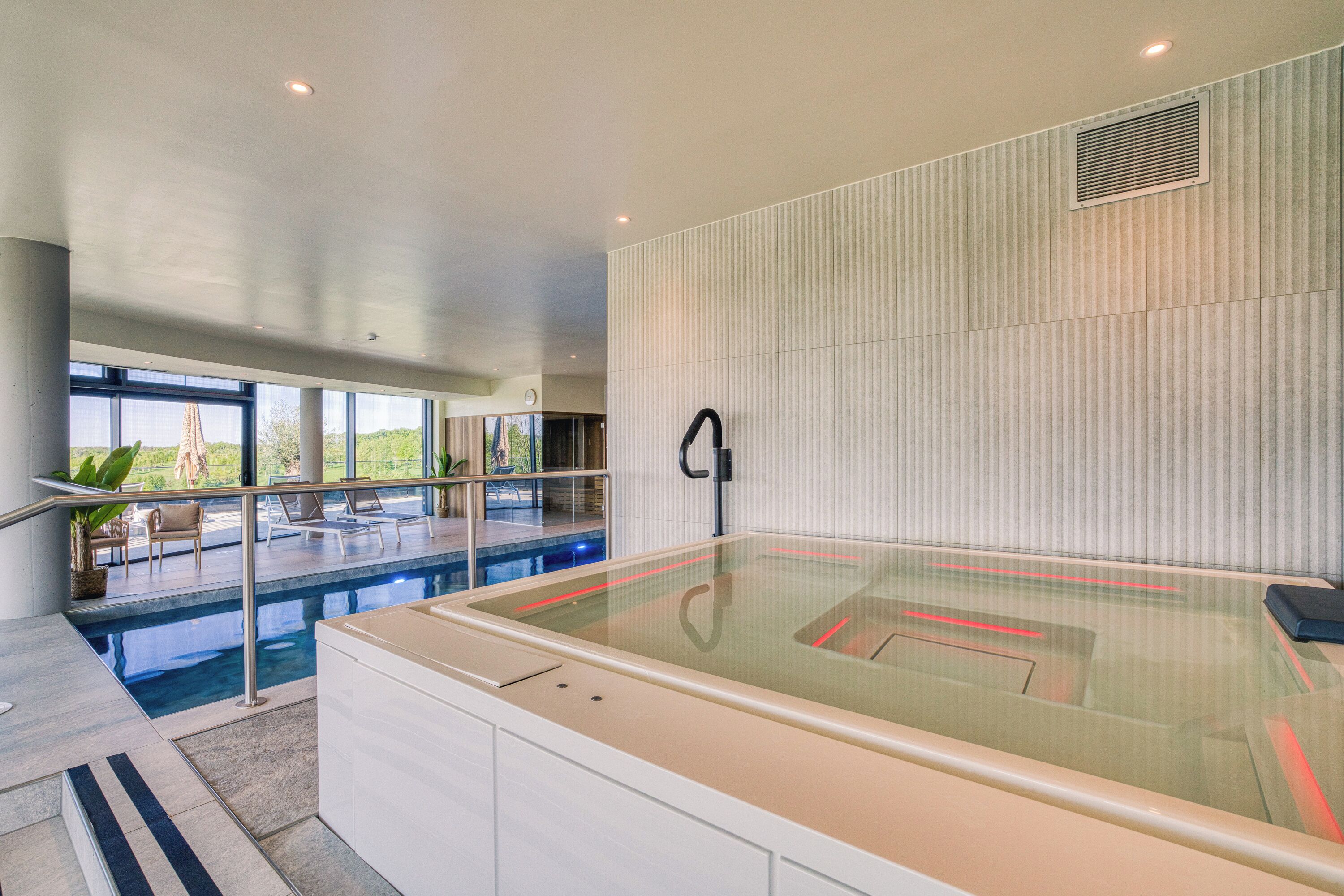A modern indoor swimming pool with a glass enclosure, a lounge area with chairs, and a view of the outdoors.
