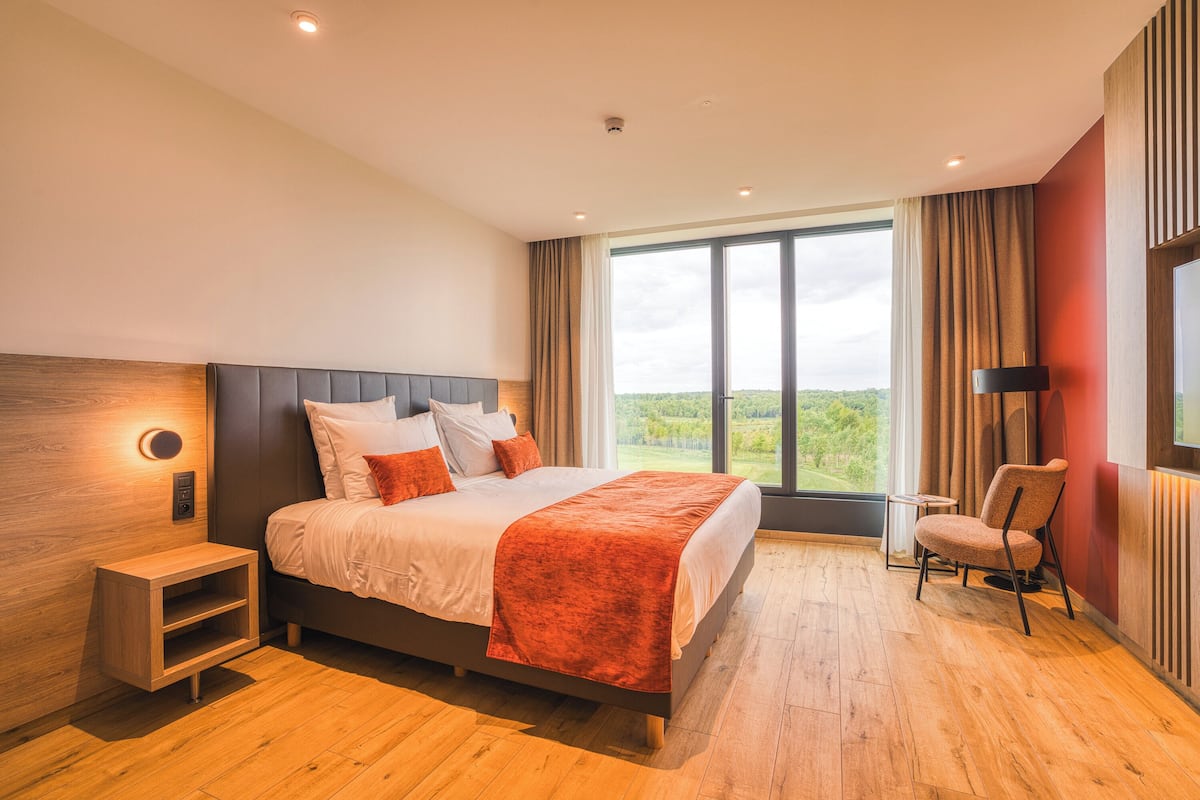 A modern bedroom with a large bed, a nightstand, a chair, and a view of the outdoors.