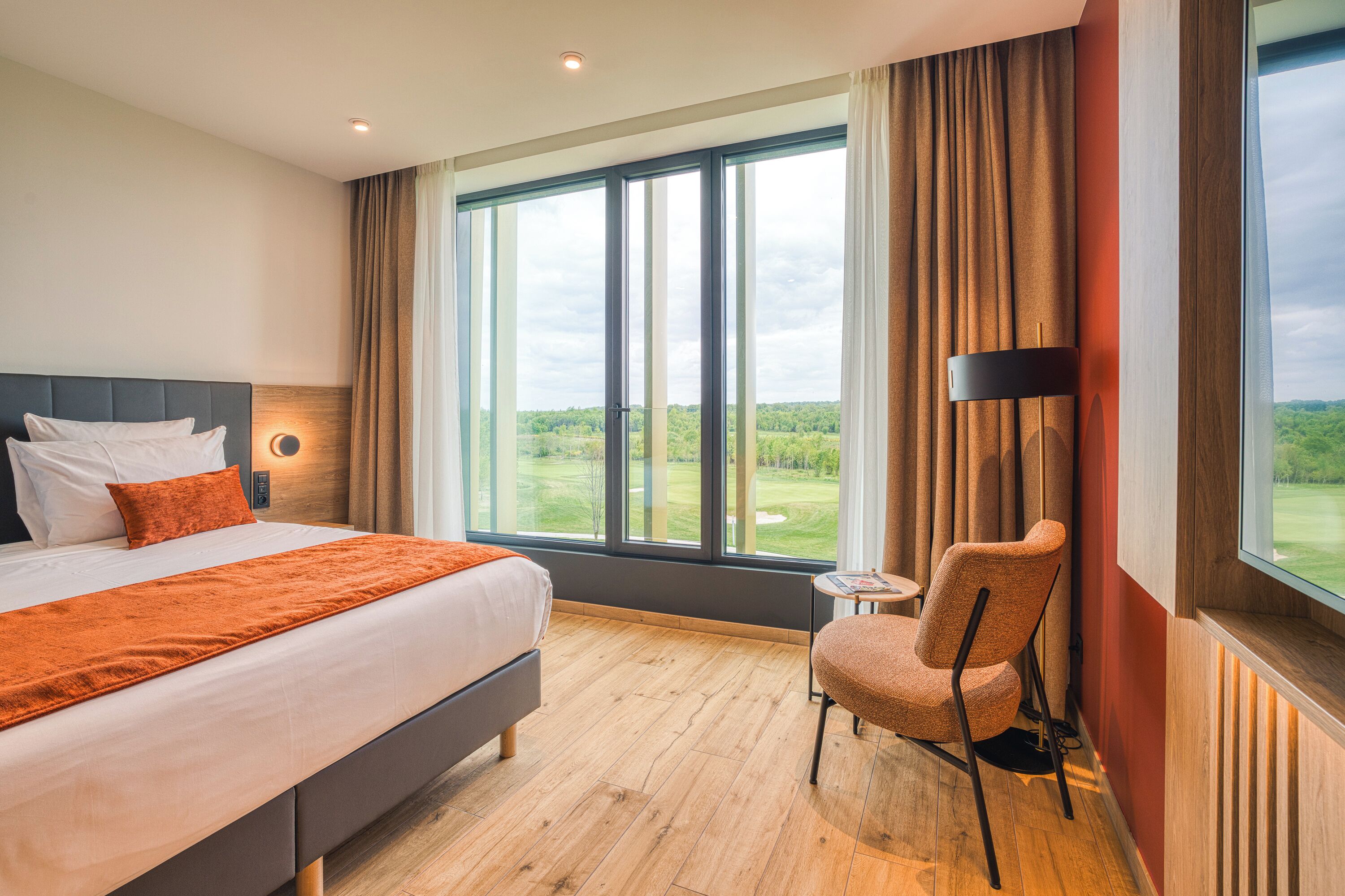 Quarto de hotel com uma cama grande, uma cadeira, uma mesa pequena e vista para um campo de golfe.