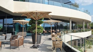 Un espace extérieur aménagé avec des meubles en osier et de grands parasols, un arbre en pot, et un bâtiment moderne aux fenêtres en verre et au design incurvé.