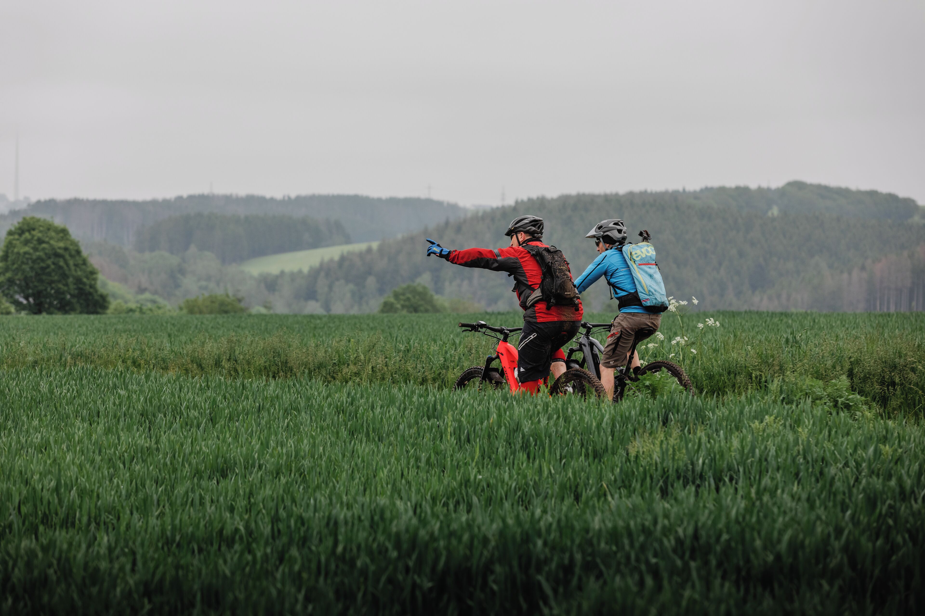 Two people on a mountain bike in a grassy field with a forested hill in the background.