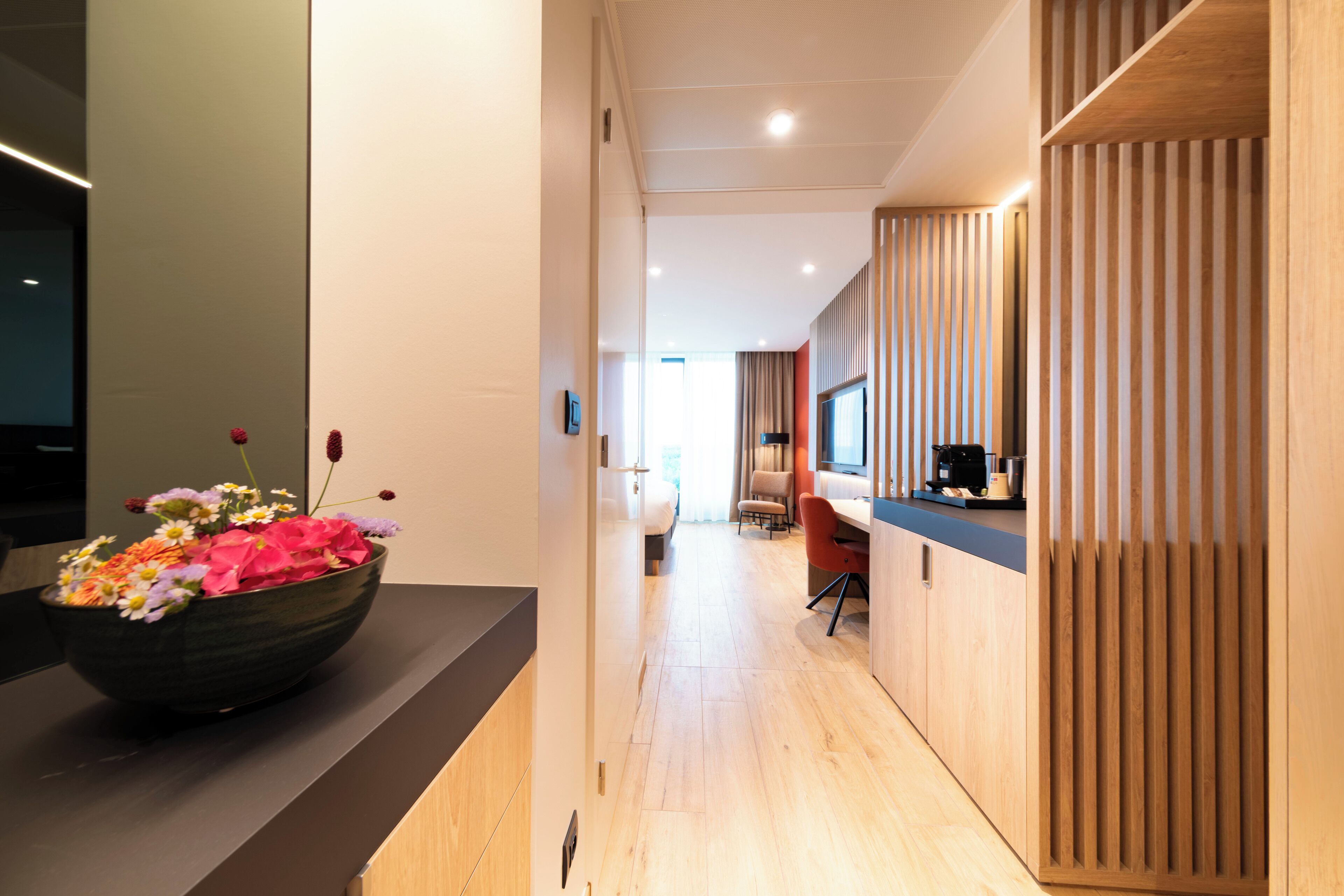 A modern hotel corridor with a wooden floor, a large black bowl of flowers, and a wooden partition on the right.