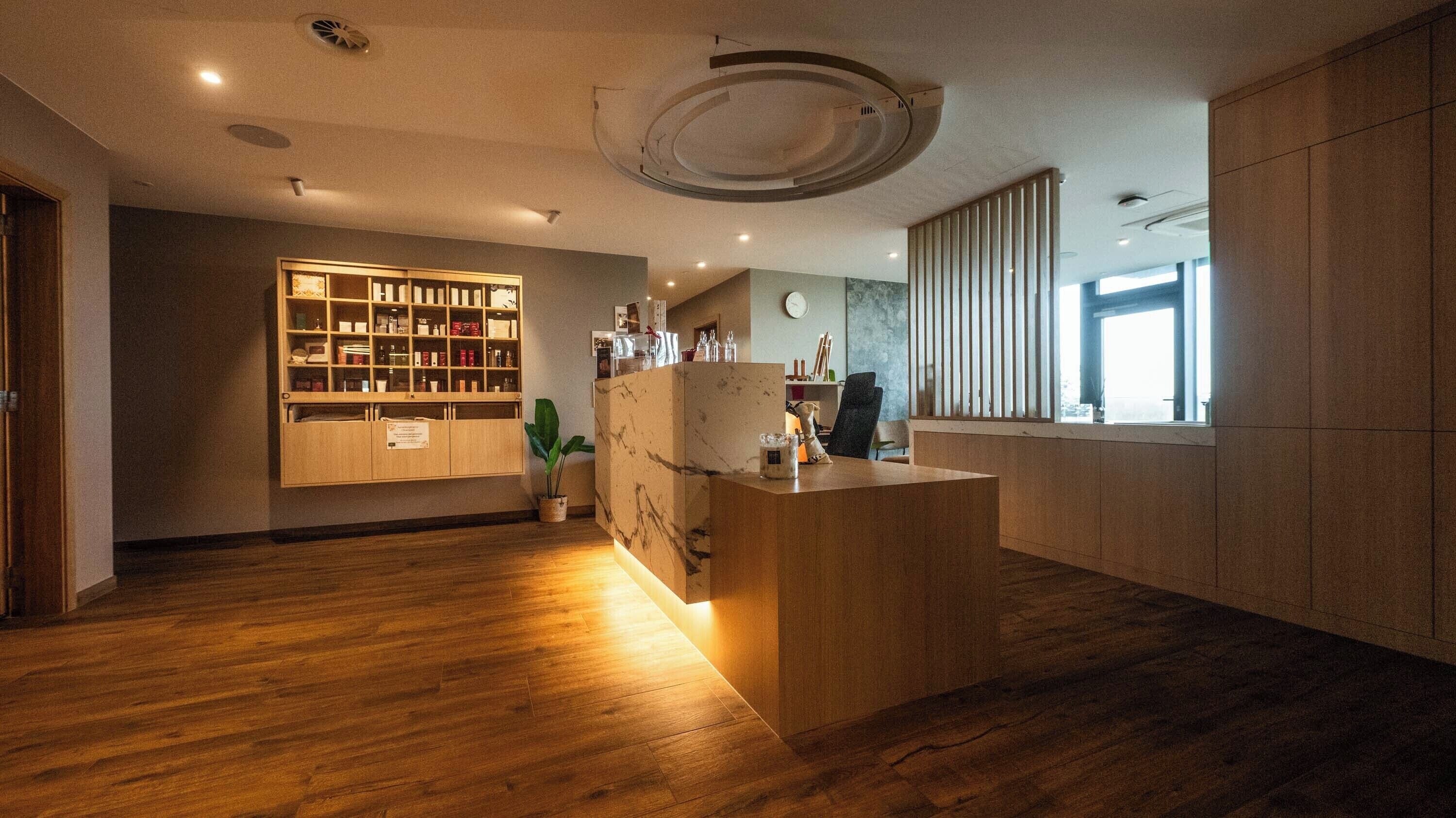 Un espace d’accueil moderne, doté d’un comptoir de réception en bois, d’une vitrine en verre présentant divers objets et d’une grande fenêtre avec des stores verticaux.
