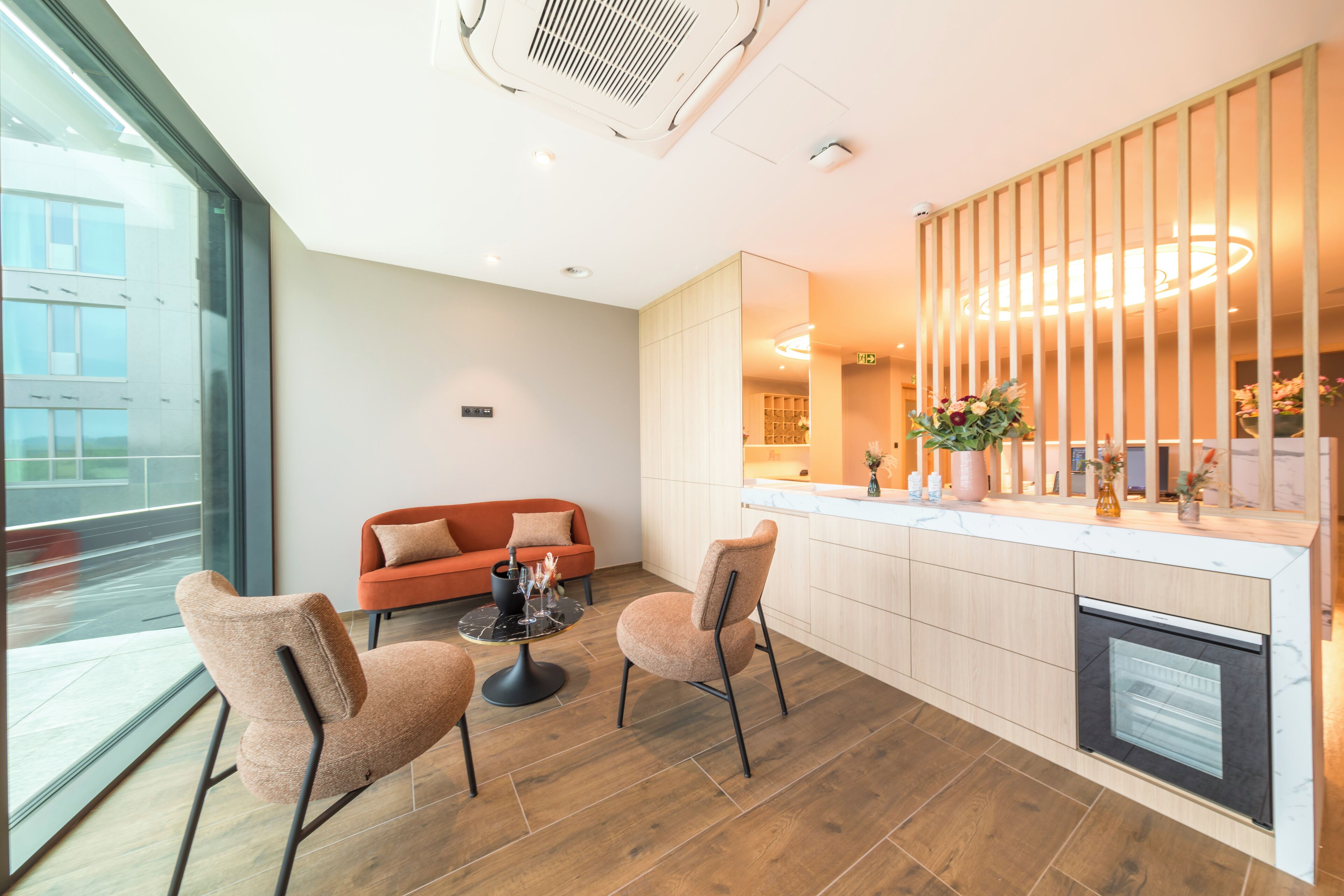 A modern hotel room with a large window, a sofa, two chairs, a small table, a kitchen area, and a decorative plant.