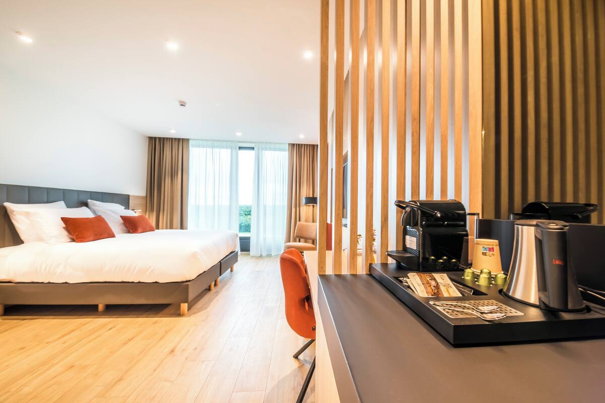 A modern hotel room with a bed, a desk with a coffee machine, and a view of the outdoors.